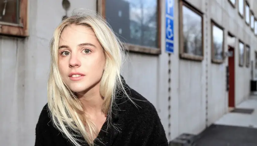 En ung kvinna med blont hår, Paolo Nobis Sandén, tittar in i kameran framför en grå byggnad med fönster.