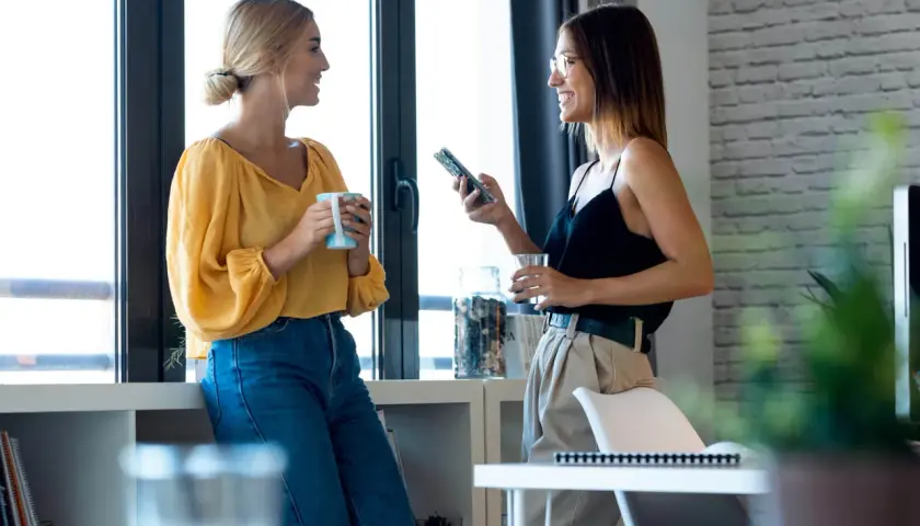 Dwie kobiety rozmawiają przy oknie, jedna z kubkiem, druga z telefonem. To przykład, jak wygląda **small talk co to** jest.