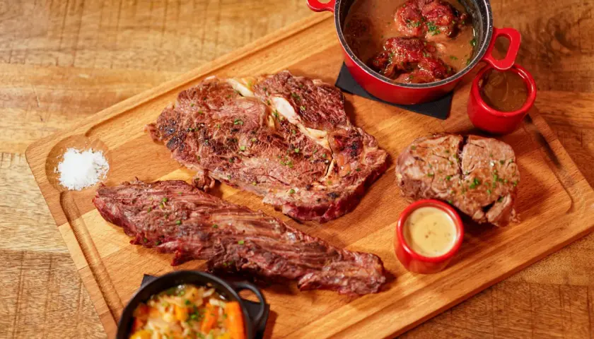 Un plateau en bois présente une entrecôte barbecue juteuse, des morceaux de viande braisée, des sauces et des légumes.