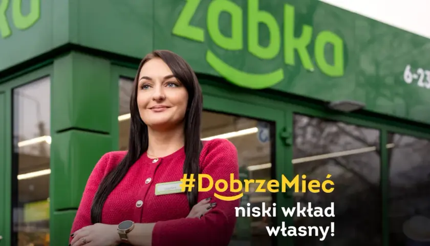 Uśmiechnięta franczyzobiorczyni Żabki przed sklepem. Hasło: #DobrzeMieć niski wkład własny!