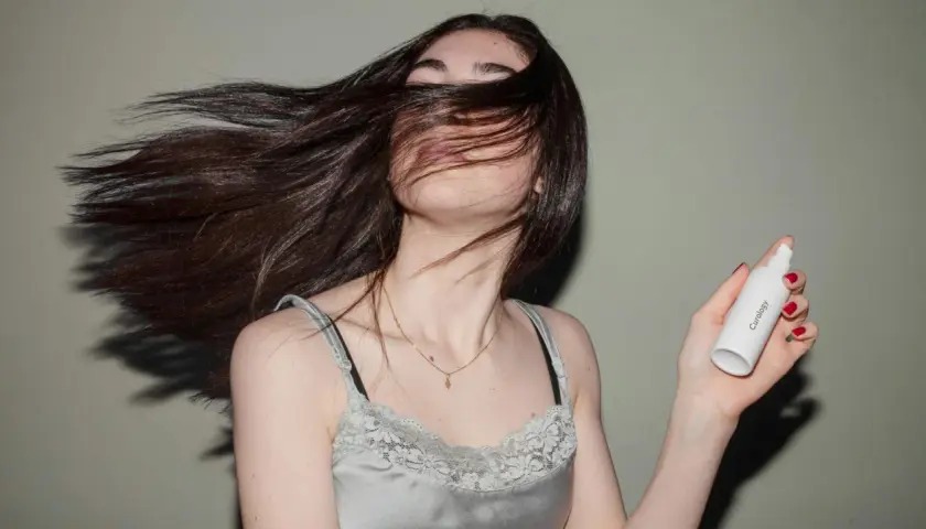 Mujer con cabello largo y oscuro moviéndose, sosteniendo un frasco de **aceite para el pelo**.
