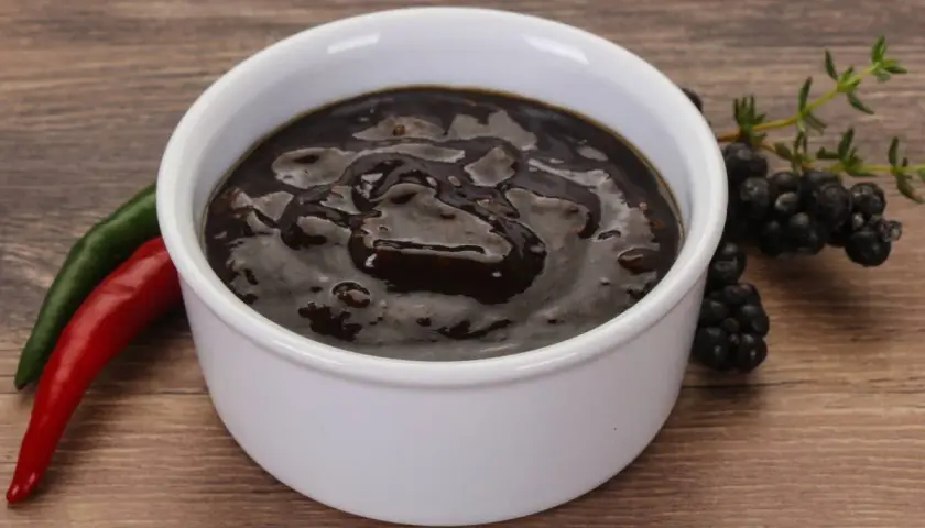 Miseczka z ciemnym sosem pieprzowym, obok papryczki chili i jeżyn.