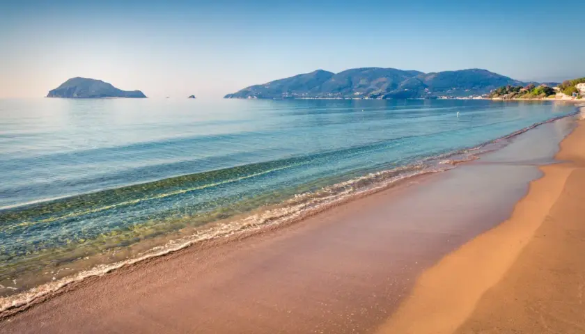 Spokojne morze obmywa piaszczystą plażę Kalamaki. W oddali widać zielone wzgórza i małe wysepki.