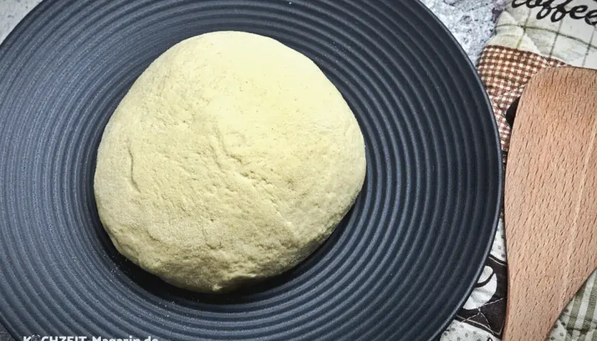 Frischer pizzateig auf schwarzer Platte, bereit für den pizzaofen. Ein Holzschaber liegt daneben.