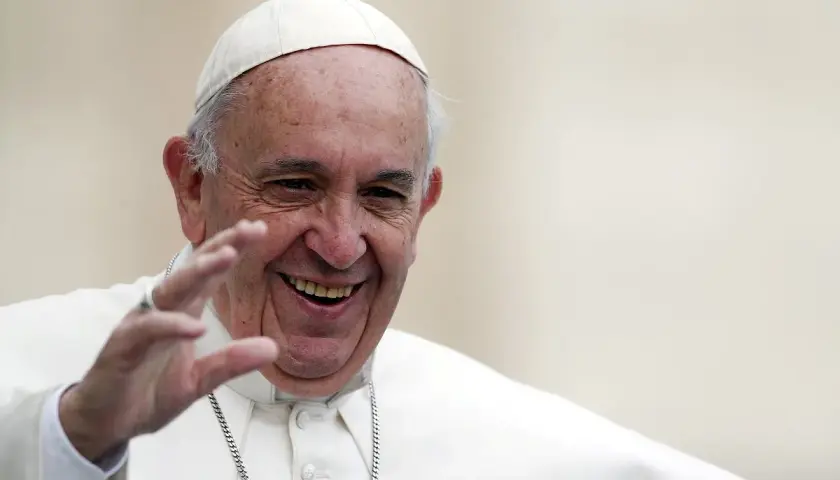Papież Franciszek w białej sutannie i piusce, uśmiechnięty, macha ręką.
