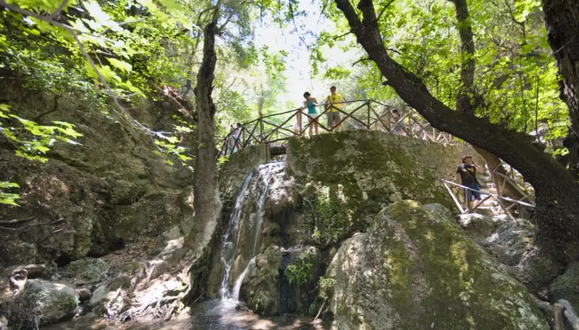 Besucher genießen die malerische Aussicht im Schmetterlingstal auf Rhodos. Ein kleiner Wasserfall plätschert über moosbewachsene Felsen, während ein Holzsteg die Wanderer durch die grüne Landschaft führt.
