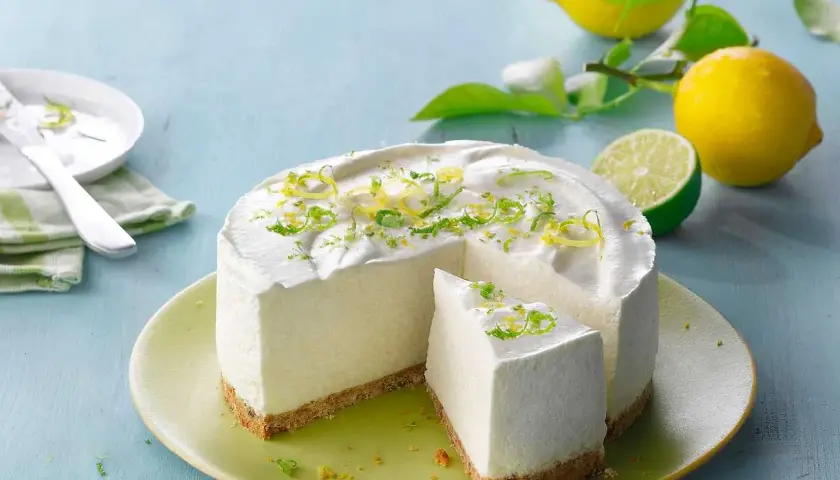En krämig citron cheesecake med rivet citronskal som dekoration. Bredvid ligger färska citroner och en lime.