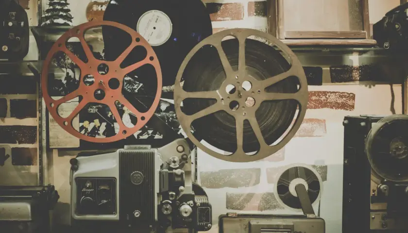 Stary projektor filmowy, taśmy filmowe i płyta winylowa na tle ceglanej ściany.