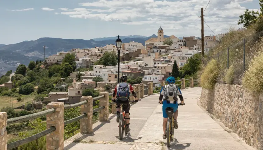 Dos ciclistas disfrutan de una ruta cicloturista ascendiendo hacia un pueblo blanco en la ladera de una montaña.