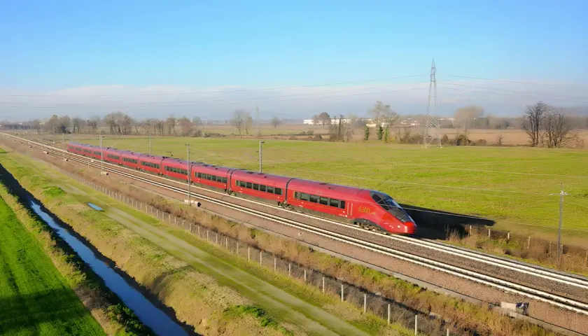Le train rouge Italo file sur les voies, reliant Roma à Naples à travers la campagne verdoyante sous un ciel bleu.
