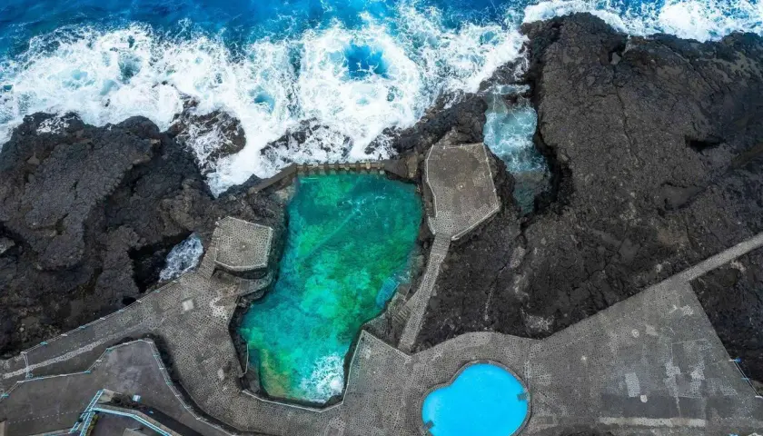Piscinas naturales de agua turquesa y azul brillante, rodeadas de rocas volcánicas y el mar embravecido.
