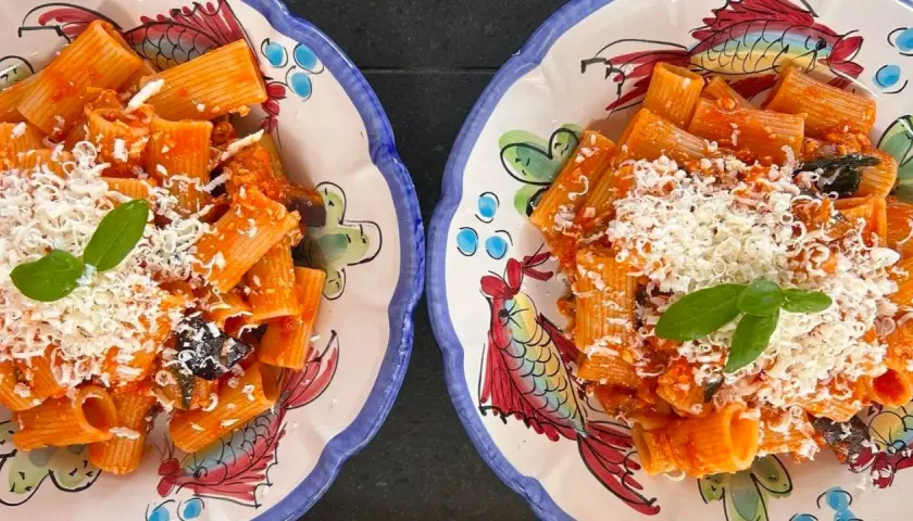 Deux assiettes de rigatoni alla norma, garnies de basilic frais et de fromage râpé, sur des plats décorés de poissons colorés.