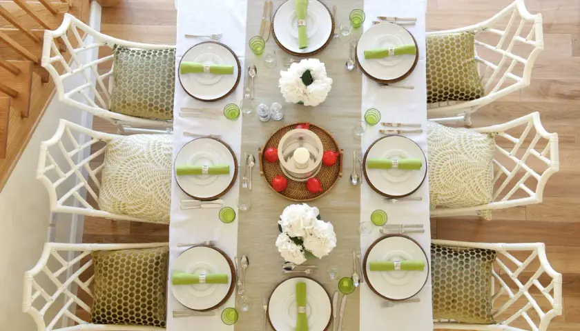 Tavola apparecchiata per una festa a tema chic, con sedie in bambù bianco e cuscini decorati.