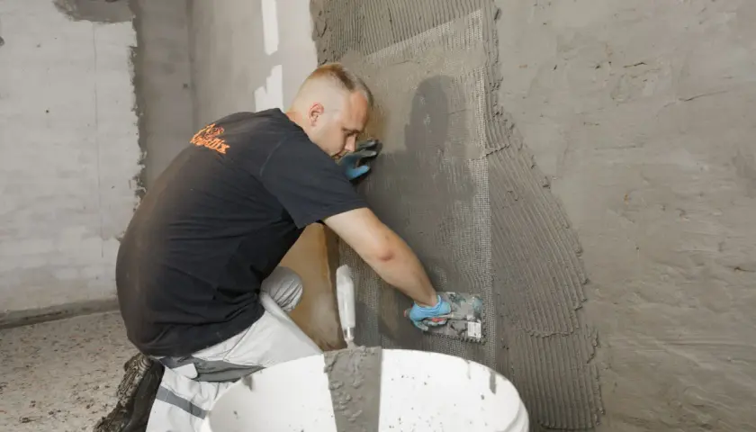 Mann trägt Mörtel auf ein Armierungsgewebe auf, um die Kellerwand von innen abzudichten.