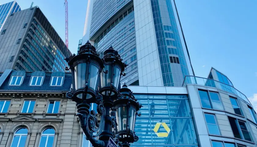 Ein historischer Laternenpfahl vor dem modernen Commerzbank-Gebäude in Frankfurt. Die Fassade spiegelt den blauen Himmel wider.