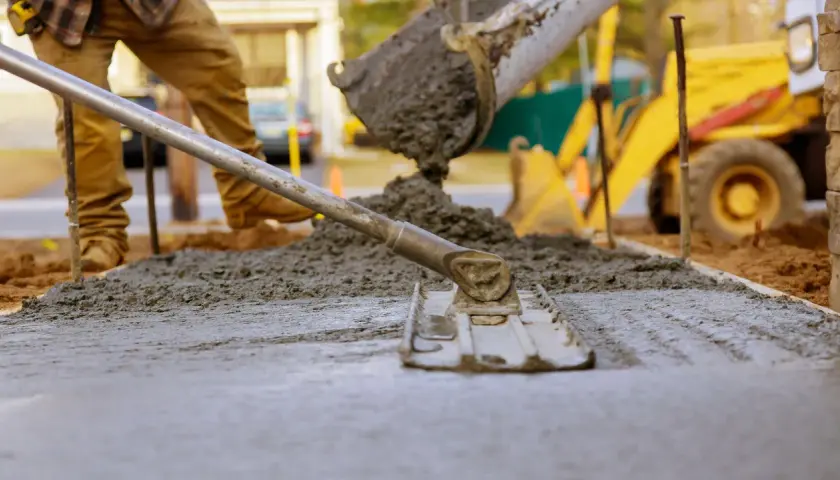 Pracownik wylewa beton na chudziaka, używając łaty do wyrównania powierzchni. W tle widać koparkę.