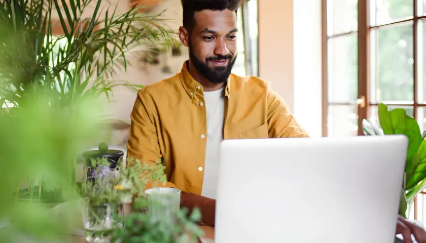 Uomo sorridente al laptop, circondato da piante. Sembra che stia imparando nuovi modi di dire per arricchire la sua comunicazione.