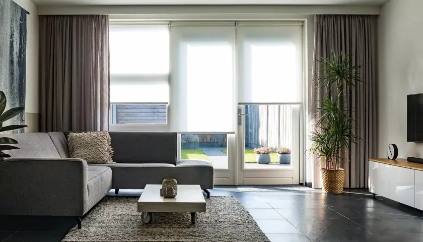 Grå soffa i vardagsrummet med draperade gardiner och stora fönster som släpper in ljus.