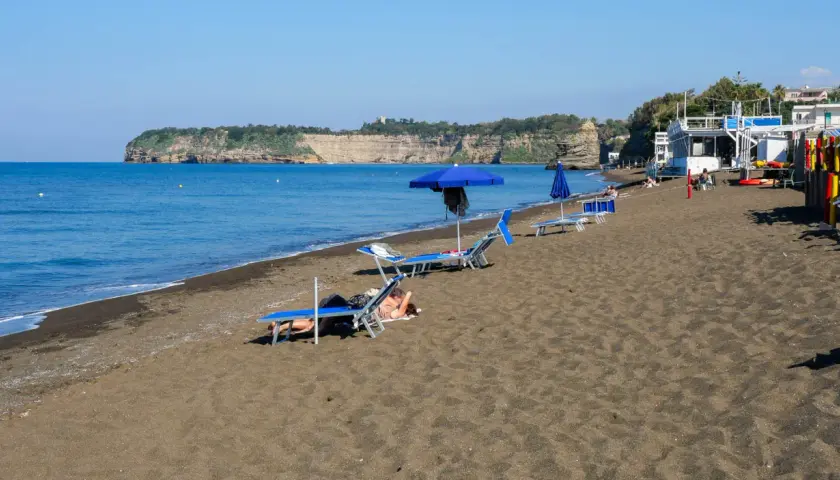 Plage de sable noir avec des chaises longues et des parasols bleus, face à une falaise rocheuse. Un doux air de Procida ou Capri.