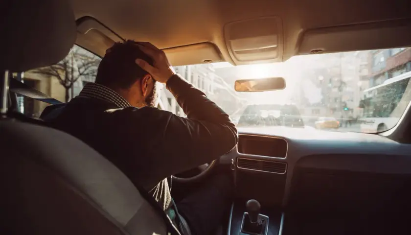 Hombre con mano en la cabeza dentro de un coche, buscando trucos para conducir sin nervios. El sol brilla a través del parabrisas.