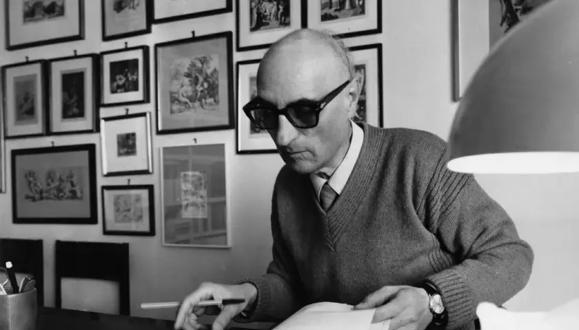 Gesualdo Bufalino, con occhiali scuri, è immerso nella lettura di un libro, circondato da quadri alle pareti.