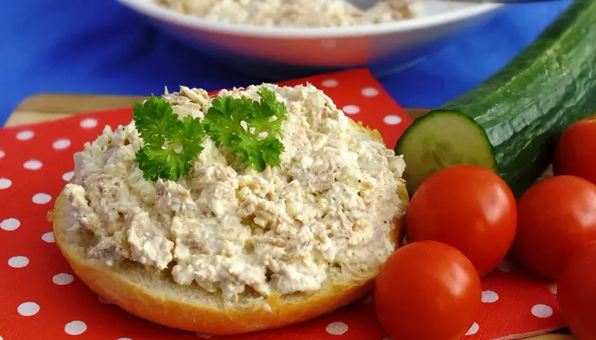 Pyszna pasta z tuńczyka i twarogu na bułce, ozdobiona natką pietruszki, obok pomidorki i ogórek.