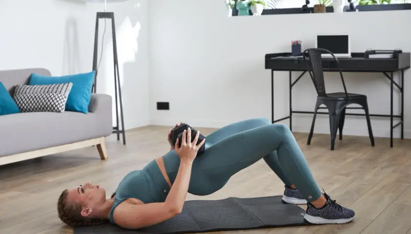 Kobieta wykonuje ćwiczenie glute bridge z obciążeniem na biodrach w domowym zaciszu.