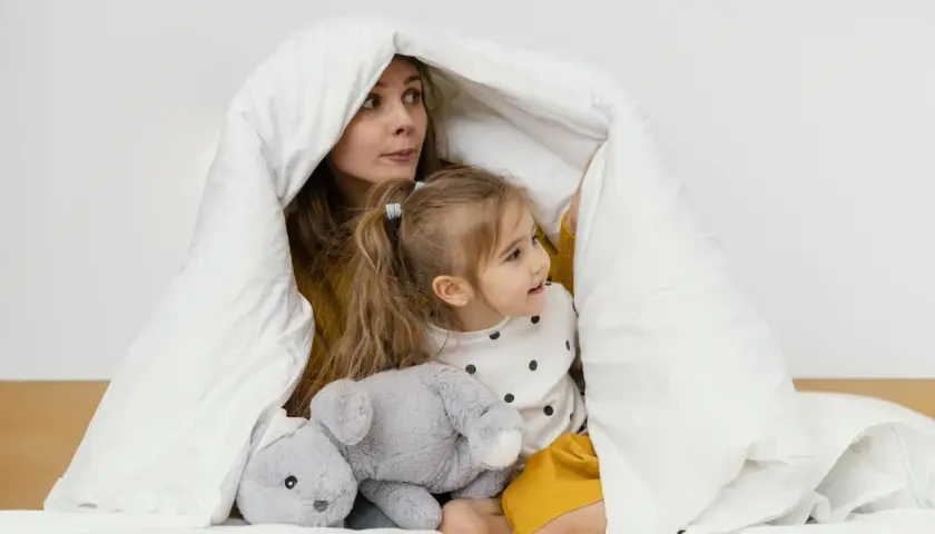 Mama i dziecko bawią się pod białą kołdrą, która przypomina przytulne kołderki dla niemowląt. Obok leży szary pluszowy króliczek.