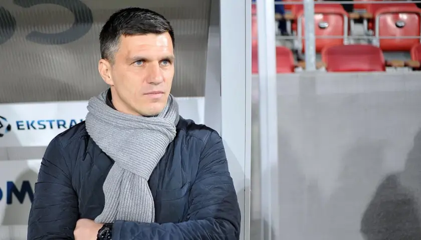 Robert Podoliński, trener w szaliku, spogląda w bok na tle stadionu z czerwonymi siedzeniami.