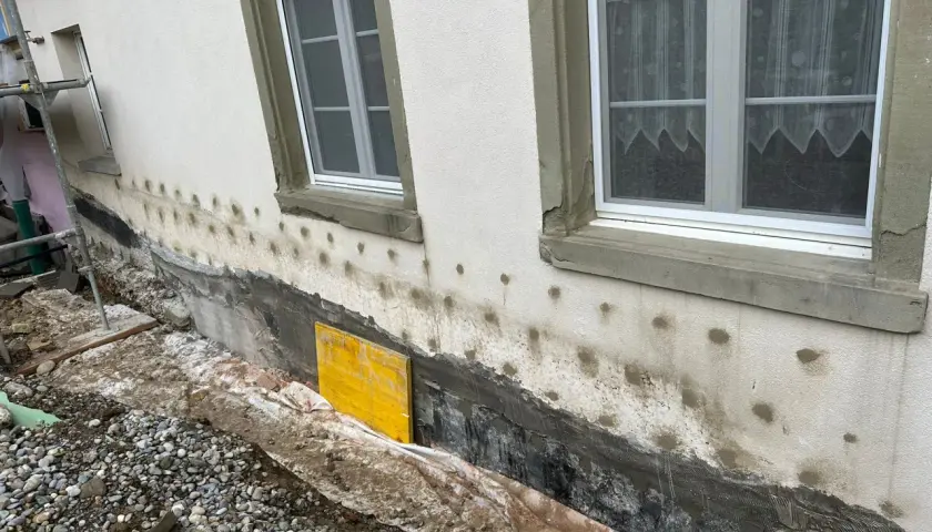 Arbeiten am Haus: Das Fundament trockenlegen mit neuer Abdichtung und Drainage. Gerüst und Baumaterialien sind sichtbar.