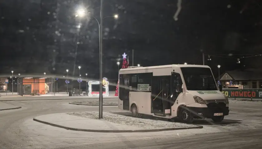 Biały bus na ośnieżonym przystanku nocą, ozdobionym świątecznymi lampkami.