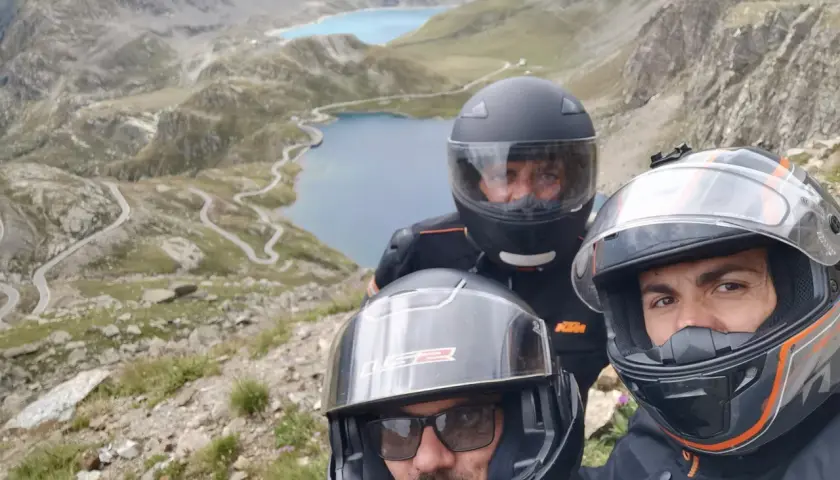 Tre motociclisti con caschi ammirano il panorama mozzafiato del Col del Nivolet, con i suoi laghi alpini e le strade tortuose.