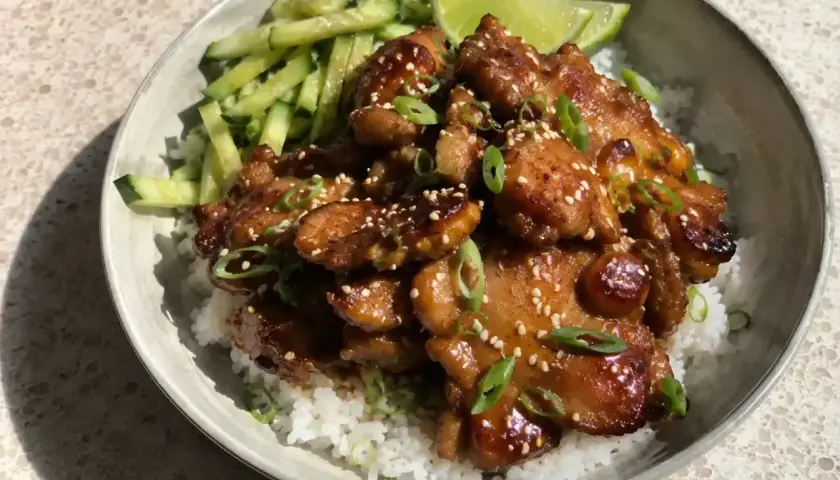 En skål med ris, gurka och saftig grillad kyckling med sesamfrön och salladslök. En limeklyfta ligger bredvid.