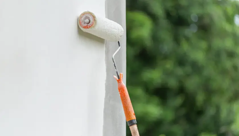 Fassade streichen ohne Reinigung: Ein Malerroller mit orangefarbenem Griff überzieht eine weiße Wand mit Farbe. Im Hintergrund verschwimmen grüne Bäume.