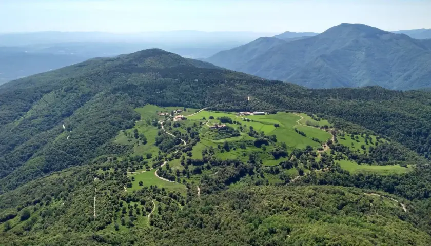 Vista aérea del **santuari del far**, rodeado de verdes colinas y bosques frondosos. Caminos serpentean entre prados y edificios dispersos.