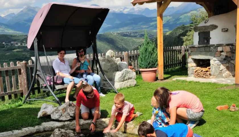 Famiglia rilassata in un giardino con vista sulle montagne, tipico delle fattorie altoatesine. Bambini giocano vicino a un laghetto, mentre adulti riposano su un'altalena.