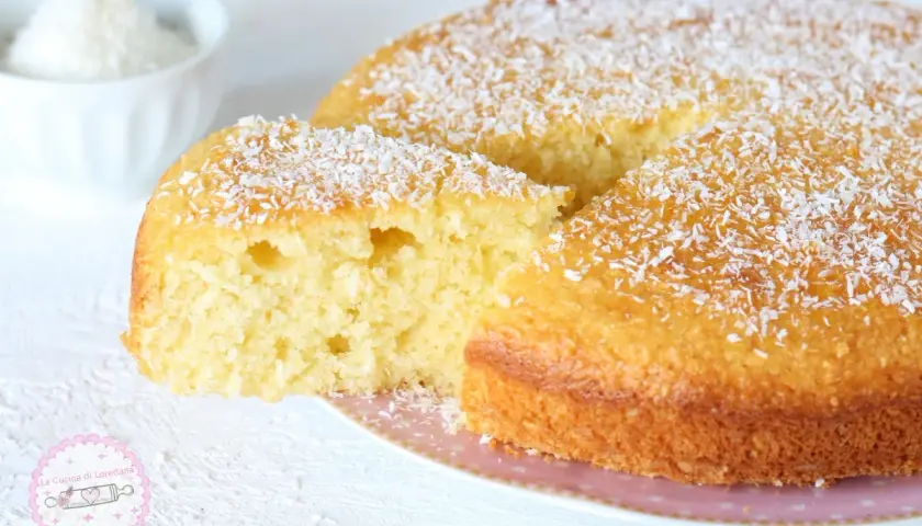 Una soffice torta al cocco, cosparsa di scaglie bianche, con una fetta tagliata che ne rivela l'interno profumato.