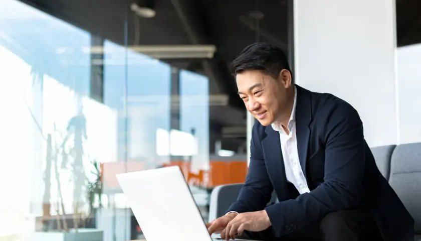 An Asian man in a suit smiles while working on his laptop, perhaps studying for his bachelors in organizational leadership.