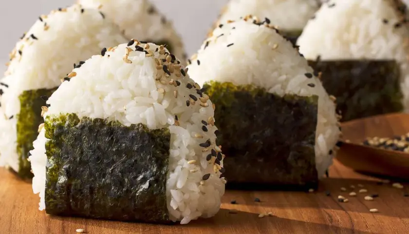 Several triangle sushi onigiri, sprinkled with sesame seeds and wrapped in nori, sit on a wooden board.