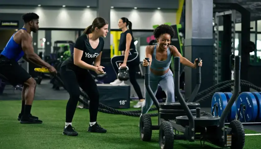 Intensywny trening interwałowy na siłowni. Kobieta pcha sanie, mężczyzna ćwiczy z linami, a inni wykonują ćwiczenia z kettlebell.