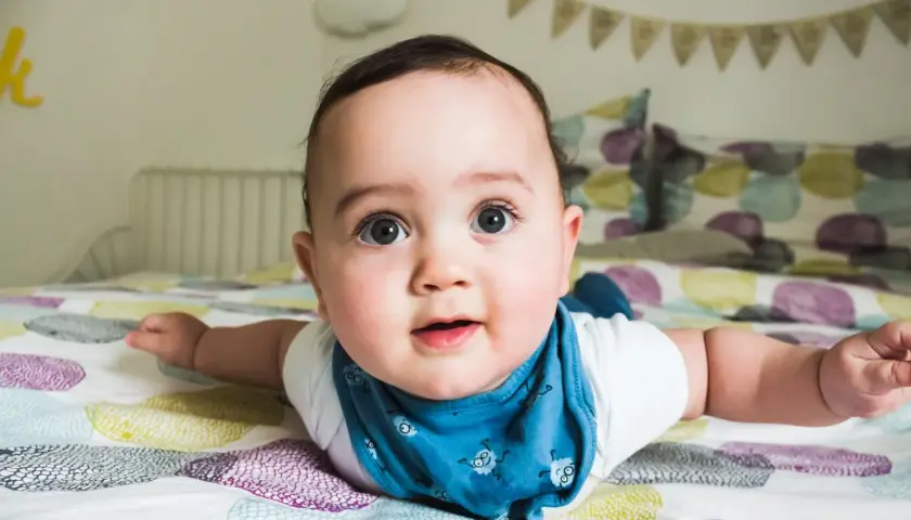 Un adorabile neonato di 4 mesi, sdraiato a pancia in giù sul letto, con gli occhi spalancati e un bavaglino blu.