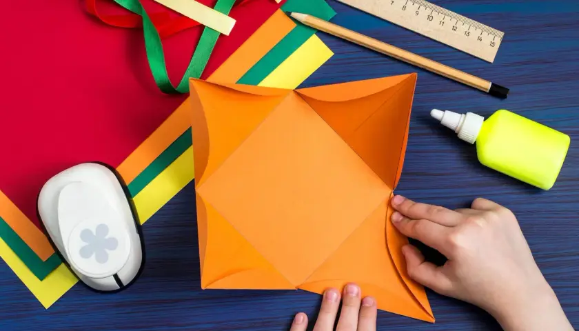 Hände falten orangefarbenes Papier für ein **Gutschein basteln**. Bastelmaterialien wie Bänder, Schere, Kleber und Lineal liegen bereit.