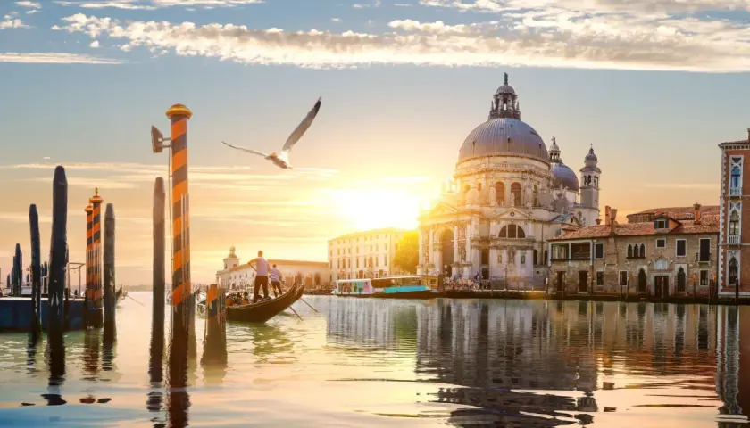 Góndola navega por canal veneciano al atardecer, con la Basílica de Santa Maria della Salute y otras **ciudades de Europa** reflejadas en el agua.