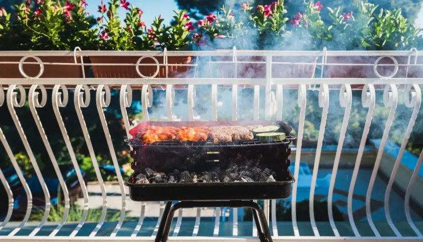 Grill z mięsem i warzywami na balkonie, w tle kwiaty i błękitne niebo.