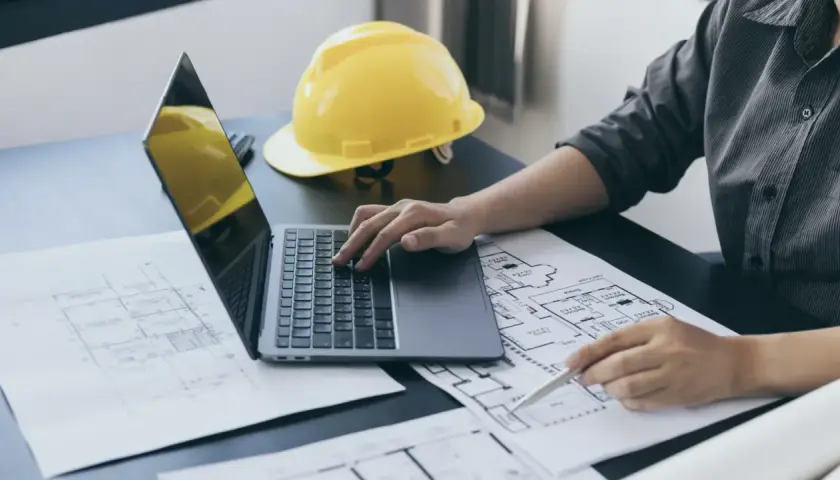 Inżynier analizuje plany budowlane, porównując powierzchnię użytkową z całkowitą. Na biurku laptop, żółty kask i dokumenty.