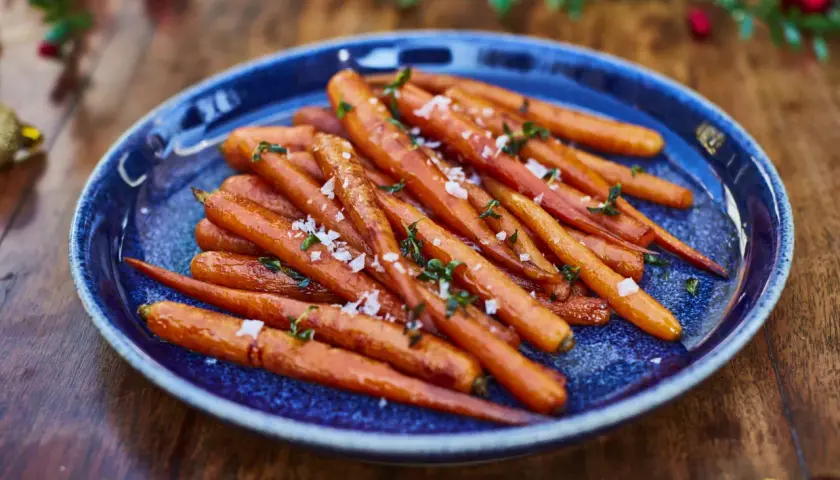 Zanahorias al horno, tiernas y doradas, espolvoreadas con sal marina y hierbas frescas, servidas en un plato azul sobre una mesa de madera.