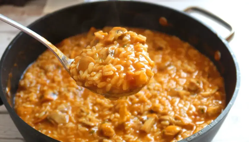 Cuchara sirviendo arroz caldoso con pollo y champiñones, humeante y apetitoso, de una sartén.