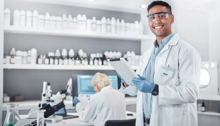 Uśmiechnięty technik farmacji w okularach ochronnych trzyma tablet w laboratorium, gdzie na półkach stoją liczne butelki, a w tle pracuje inna osoba.