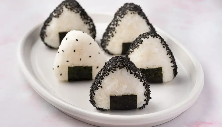Five triangular rice balls, some coated in black sesame seeds, sit on a white plate. This is what a rice ball looks like.