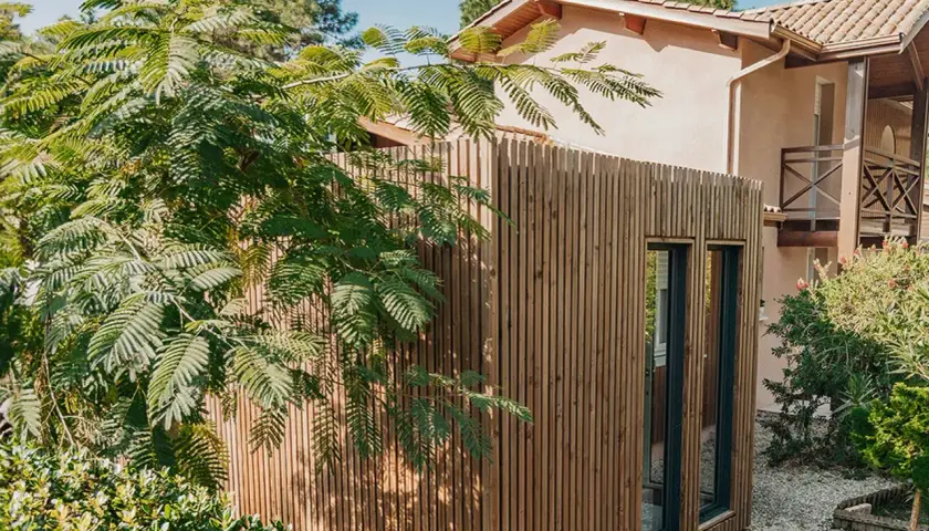 Extension de maison en bois moderne, bardage vertical, fenêtres sombres. Un espace de vie supplémentaire sans permis de construire, intégré dans un jardin verdoyant.